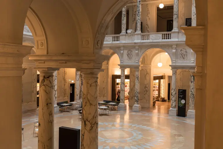 Eine großzügige Innenansicht eines Gebäudes mit hohen, marmornen Säulen und einer hellen, einladenden Atmosphäre. Im Hintergrund sind Regale zu sehen, die Teil einer Galerie oder Bibliothek sein könnten. Sanfte, runde Lichtquellen hängen von der Decke und illuminieren den Raum.