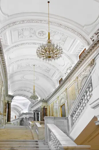 Eine elegante Treppe in einem prachtvollen Innenraum mit hohen, kunstvoll verzierten Decken. Kristallleuchter hängen von der Decke, und große Fenster sorgen für natürliches Licht. Die Wände sind mit aufwendigen Verzierungen und Gemälden geschmückt.