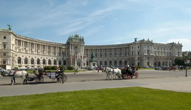 Pferdekutschen fahren vor einer historischen Architektur in einer großen, grünen Fläche. Im Hintergrund ist ein beeindruckendes Gebäude zu sehen. Der Himmel ist klar und blau.