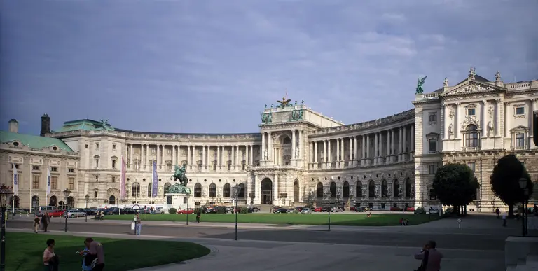 Ein breiter Blick auf das historische Gebäudeensemble des Wiener Hofburg mit einer großen Rasenfläche im Vordergrund. Der Himmel ist bewölkt, und Menschen bewegen sich im Gelände. Die Architektur zeigt prächtige Fassaden und eine Statue in der Mitte.